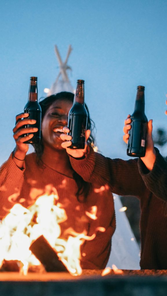 Group of friends celebrating with drinks around a cozy campfire during sunset.